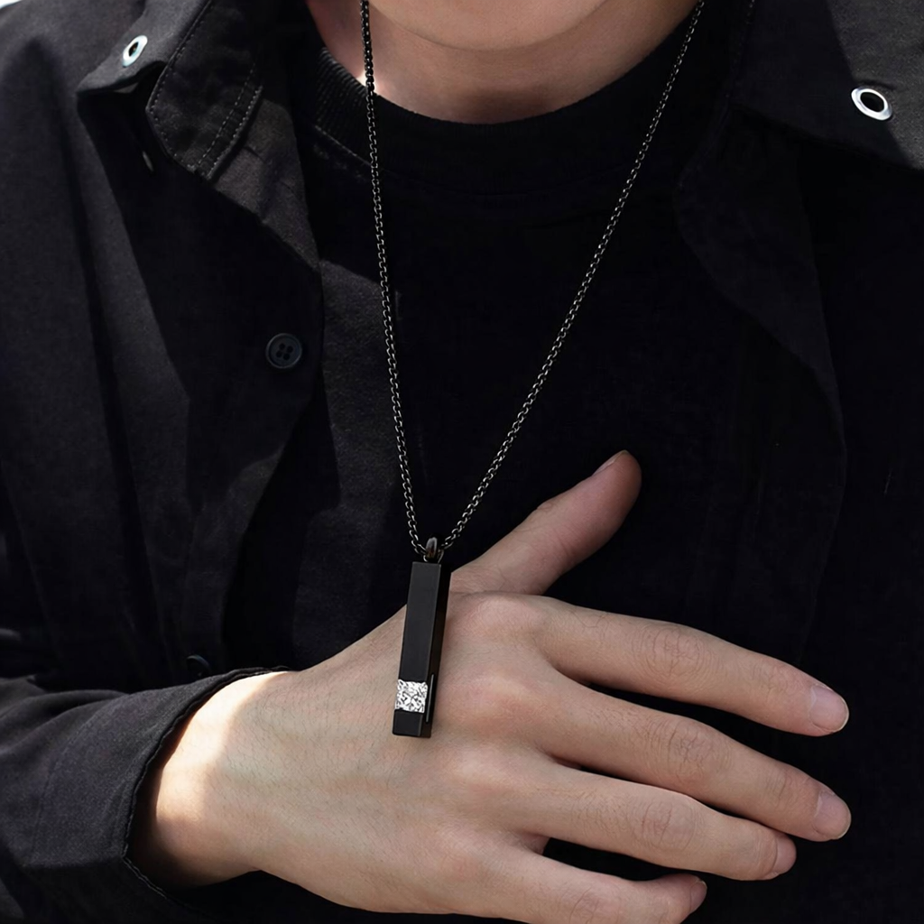 Person wearing a black pendant necklace with a diamond, held in front of a black shirt.